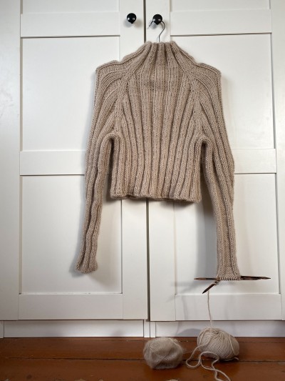 Beige Midnight jumper hanging on a wardrobe door with one sleeve still being knitted on needles.