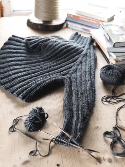 Dark grey Midnight jumper knitting pattern shown in progress on a wooden table with one sleeve on needles.