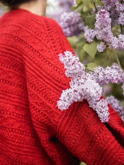 Back detail of the red Ruke Moon cardigan knitting pattern beside lilac flowers