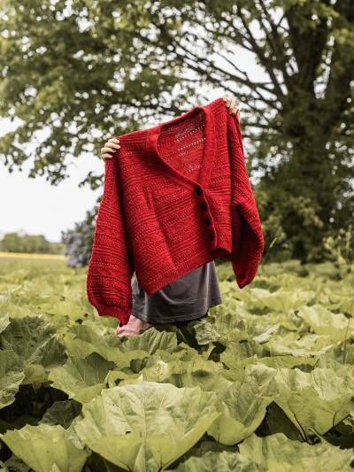 Red Ruke Moon cardigan knitting pattern held up outdoors above green leaves